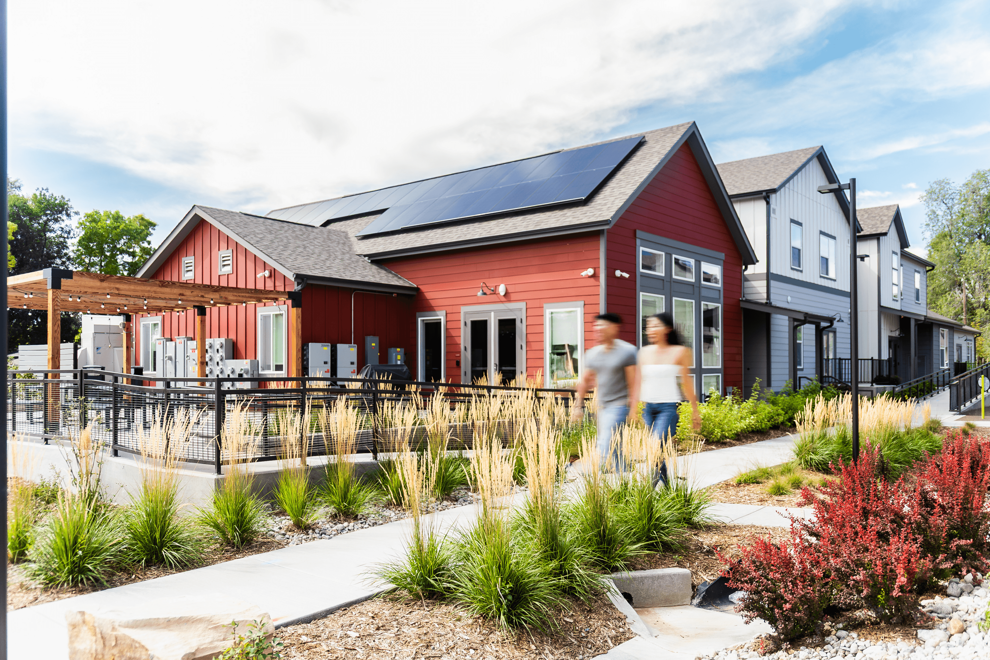 A red building with solar panels on the roof is surrounded by a garden with people walking.